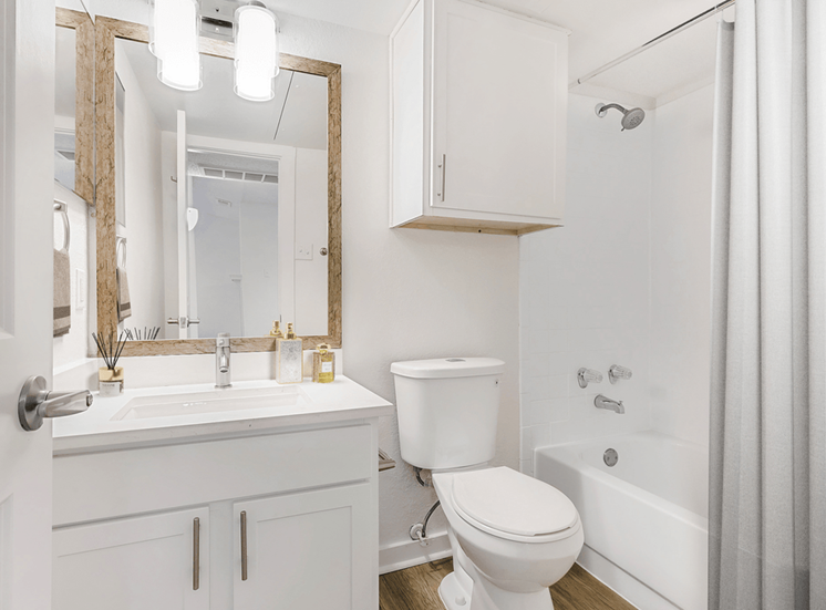 Virtually staged bathroom with white cabinetry, white quartz countertop, custom vanity mirror, medicine cabinet, wood style flooring, sheer shower curtain and toilet
